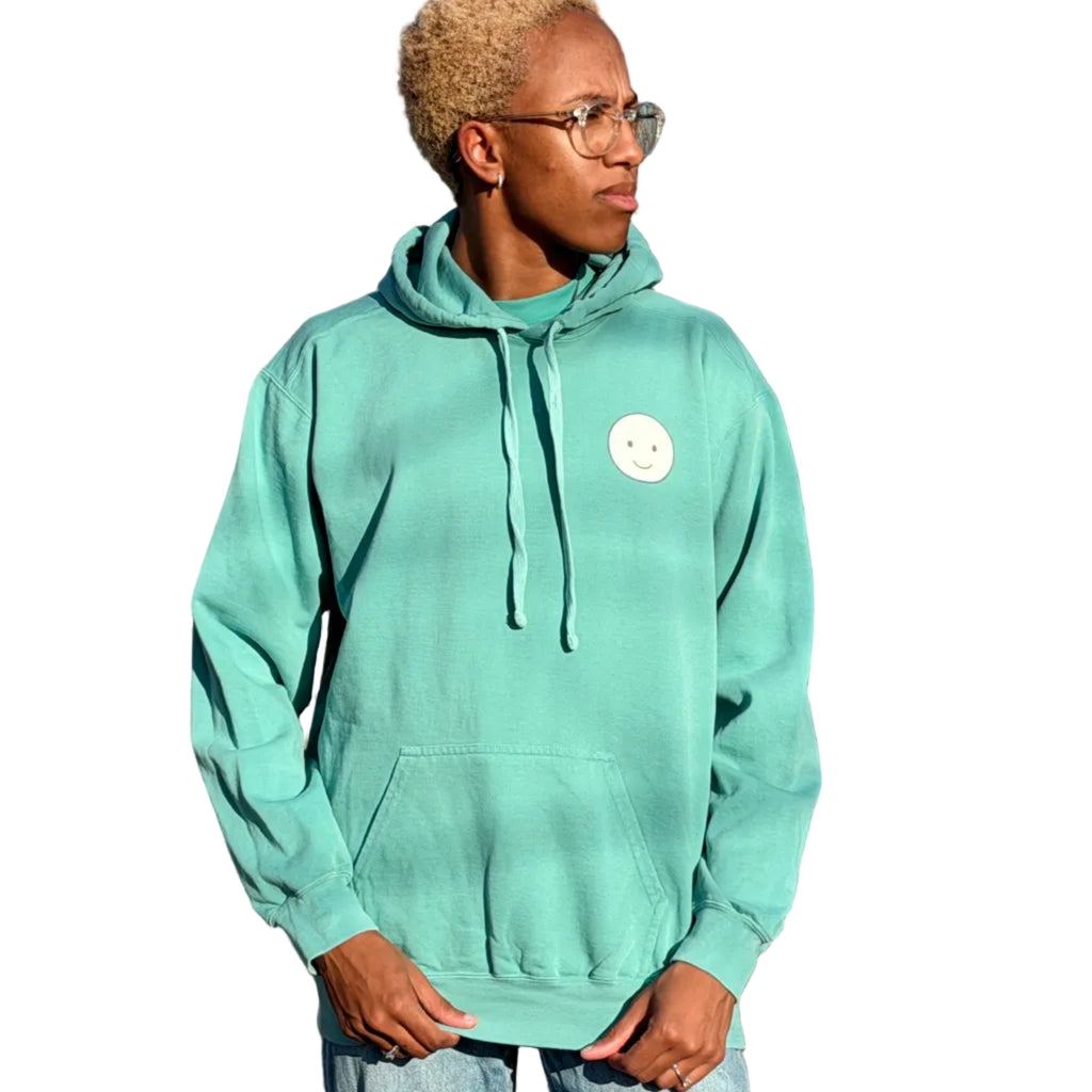 Person wearing a light blue hoodie with a logo, standing in an outdoor setting with trees and a clear sky.