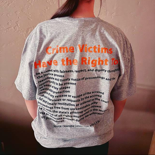Person wearing a gray t-shirt with text on the back, standing against a plain wall.