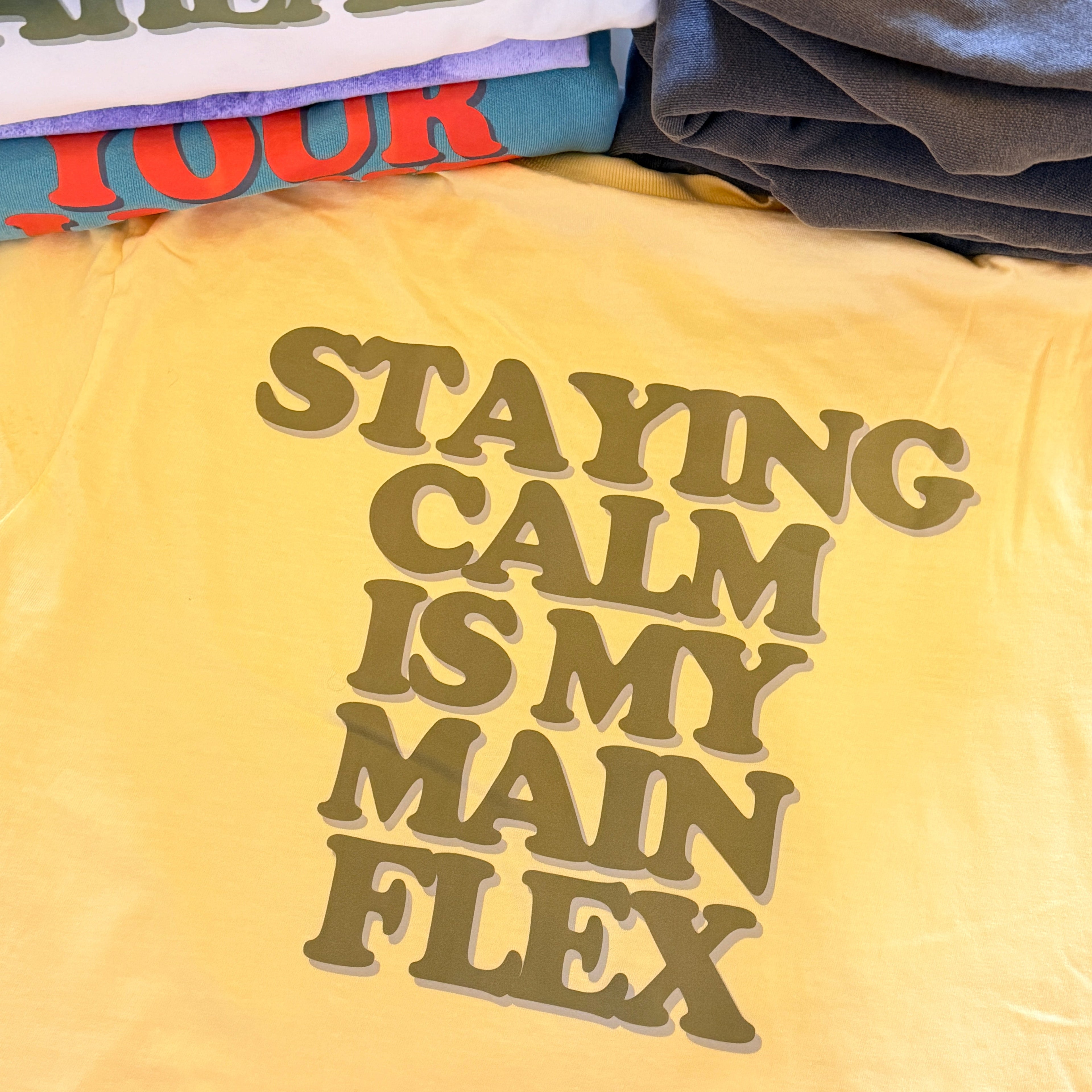 Yellow t-shirt back with text 'STAYING CALM IS MY MAIN FLEX' on a table with other First Light items.