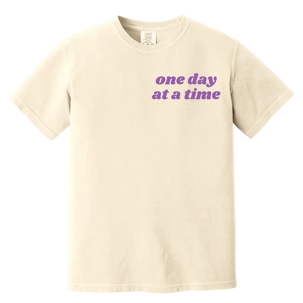 Beige t-shirt with 'one day at a time' text in purple on a white background